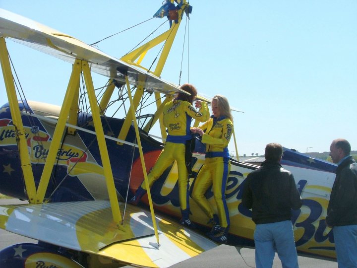 Amazingly Pistonheads Aircraft - The image showcases a lively scene at an airshow. A vintage airplane, painted in shades of blue, yellow, and white, is the main attraction. Its propeller, blades turned, is ready for flight. Two young individuals, dressed in yellow flight suits, are standing next to the airplane. They appear to be preparing to climb aboard, perhaps for a demonstration or flight. Two spectators are situated on the runway, meandering near the airplane, possibly discussing the upcoming events or simply admiring the aircraft. The sky above is a clear blue, providing a serene backdrop to the bustling activity on the ground. The overall atmosphere exudes the excitement and anticipation typically associated with such aviation events.