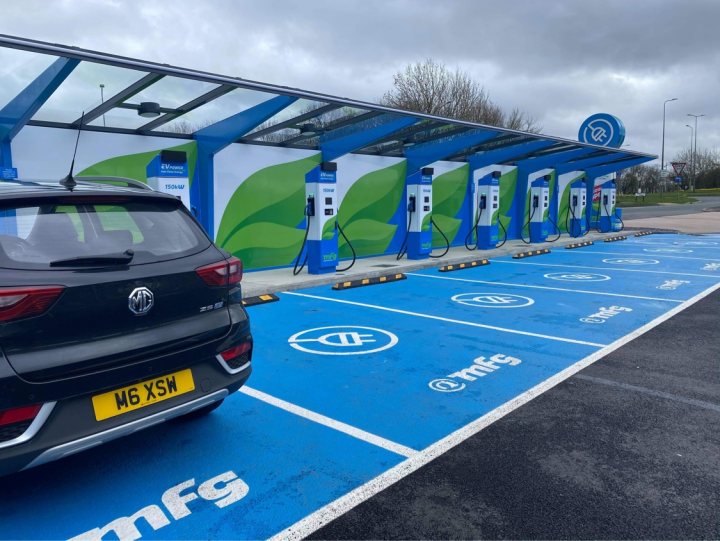 Rolling the electrified dice. 2020 MG ZS EV Exclusive  - Page 8 - Readers' Cars - PistonHeads UK - The image shows an electric charging station with a blue color theme. The station is equipped with several charging points and has a sign displaying information about the chargers, such as payment options. A dark-colored car is parked at one of the charging stations, indicating that it might be in the process of charging. The surrounding environment includes a road adjacent to a parking area, and there are trees and buildings in the background, suggesting an urban setting.