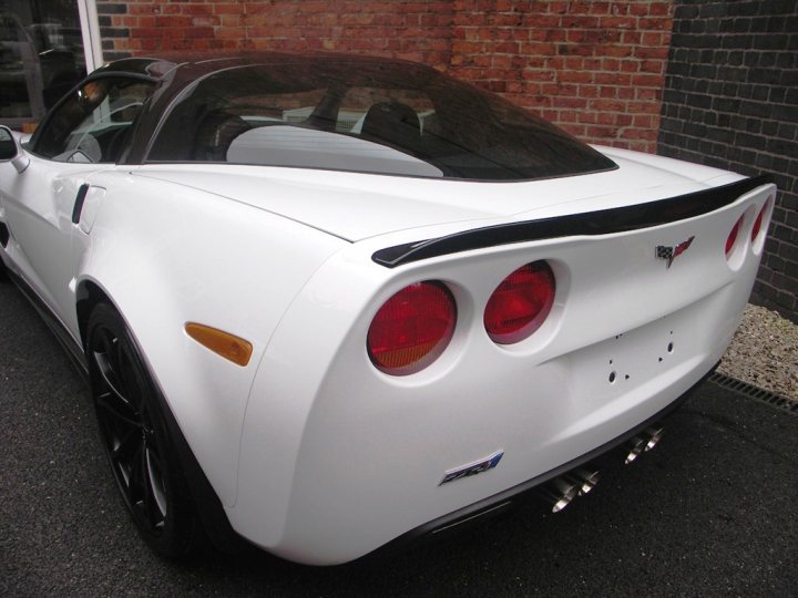 How about some ZR-1 photos to cheer you up? - Page 1 - Corvettes - PistonHeads - The image features a white sports convertible car parked on a paved surface outside a brick building. The vehicle is sleek with black trim accents, and a distinctive emblem is visible on the upper right side of the rear fender. The license plate is centered and white, with visible model-specific details and numbers. The setting appears to be a residential or commercial area, given the presence of a brick wall and a paved driveway.