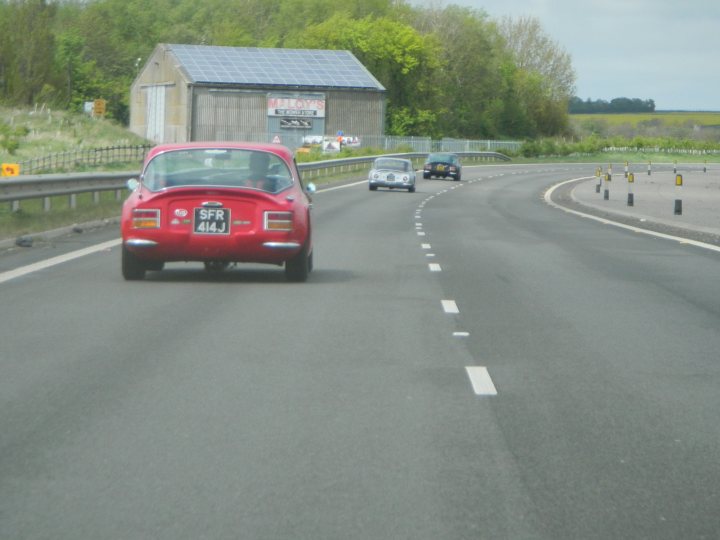 Early TVR Pictures - Page 80 - Classics - PistonHeads - The image shows a three-lane highway with a red car and a white car in the middle lane, traveling in the same direction. The lighting suggests that it's daytime. On the right side of the road, there's a curved section leading to a toll booth or speed control station in the distance. The toll booth appears to be a two-lane structure. The vehicles have released emissions, indicating petrol engines, and there are traffic markings indicating a fast lane. The sky is overcast.