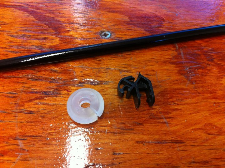 Pistonheads - This image shows a tabletop with a few scattered items. Prominently featured are a black and silver binder clip, a small white plastic disc, and a dark metal key. The binder clip has curved metal bars holding the binding tabs in place. The plastic disc has a hole in the center and appears to be a spool with some material partially rolled up. The key has a standard circular head supported by a flat and elongated blade. The tabletop has a rough, woodgrain surface texture.