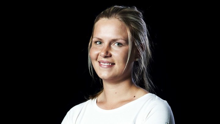 A woman in a white shirt and black tie - Pistonheads - The image shows a woman with fair skin and blonde hair, which appears to be styled upwards, possibly in a bun or updo. She has two strands of hair that have fallen down in front of her face. The woman's facial expression is warm and friendly, with a smile that showcases her teeth and lips. She is wearing a white top that stands out against the black background. The background is uniformly dark, highlighting the woman as the central subject.