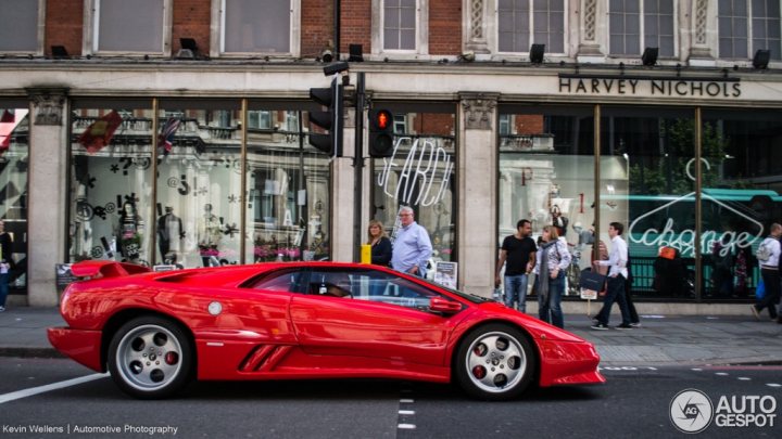 V12 Lambo Photo Thread - Page 3 - Diablo/Murcielago/Aventador - PistonHeads - The image is a bustling city scene with a distinctive red sports car prominently in the foreground. The sports car is stopped at a traffic light, under which it has just passed. The traffic light is attached to a street light at an intersection. The sports car has a sleek design with a large rear wing and alloy wheels. In the background, a crowd of people is visible on the sidewalk, and various shops can be seen, suggesting a commercial area.