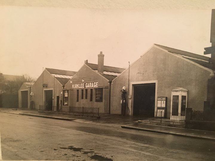 Glasgow car dealers in the 60's -70's - Page 16 - Scotland - PistonHeads - The image is a black and white photograph of a street from what appears to be a past era, given the style of the cars, people, and the architecture of the buildings. The buildings, possibly old garages, stand on the corner of a street, suggesting a urban environment. Several cars and people can be seen, some in the distance and some closer to the viewer. The sky is overcast, contributing to the vintage feel of the image.