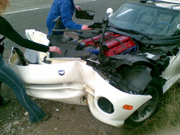 Repairing a GTS Front Fasica  - Page 1 - Vipers - PistonHeads - The image captures a scene of two individuals examining a white vehicle with an open engine hood. The car appears to be slightly damaged, with the front fender bent and the hood up. Various tools and parts, including a large engine cover, are scattered around the vehicle. It seems the scene could be that of an accident or repair situation.