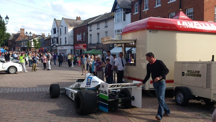 Ormskirk MotorFest - Page 1 - North West - PistonHeads - The image depicts an outdoor scene of a vehicle display. There's a man, dressed in a black t-shirt and jeans, standing next to a beige and white vehicle. He is in the process of opening the hood of the car. The car is positioned on a street corner, with buildings visible in the background. Several onlookers are gathered around, observing the man's actions with curiosity. Another vehicle is parked in the background and there appears to be a building with a red canopy. The overall setting suggests an event or exhibition where the public is allowed to interact with the displayed vehicles.