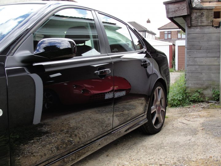 Pistonheads Newbiewell Saab - The image features the passenger side of a gray car parked on a gray, gravelly surface. The reflection in the car's door reveals a red cover that appears to be protecting something underneath. The setting includes a clear sky, suggesting a calm weather condition, and a wooden structure with a red door is partially visible in the background. The clear reflection on the car's surface enhances the contrast between the gray and red colors, creating a visually interesting image.