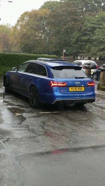 STOLEN - Blue Audi RS6 - YL15VDA - Page 1 - General Gassing - PistonHeads - The image shows a blue Audi A7 parked on a wet, road-like surface during the daytime. The car is positioned at an angle, facing the left side of the image, and appears to be parked in a lot amongst other vehicles, some of which are barely visible. The trees in the background, along with the wet conditions, suggest a park or a well-maintained city street. There is no visible text on the image.