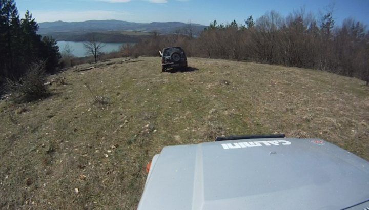 Bit of off-roading, and a bit of camping. - Page 1 - Off Road - PistonHeads - The image shows a car parked on a grassy hillside in a natural setting. The car is facing away from the camera and seems to be positioned on a path or near the edge of the road. The landscape around the parking area is filled with grass, shrubs, and a few trees, suggesting a rural or semi-rural location. In the background, there is a body of water, which could be a lake or reservoir, and distant hills or mountains providing a scenic view. The setting appears to be serene and peaceful, with minimal human activity other than the parked car.