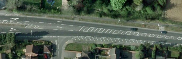 HGV Drivers. Dodgy overtaking. What do you think? - Page 3 - Advanced Driving - PistonHeads - The image presents an aerial view of a curved, elevated road circling around parked cars. Farther along the route, a pair of vehicles are visible, one of which appears to be a car, given its shape and size in relation to the segments of the road seen closest to the front of the image. The surrounding areas show a mix of greenery and urban structures. A significant part of the road seems to be elevated above a level where there are no cars either parked or moving. Finally, in the far distance, there is a small structure that stands out against the open space, suggesting a rural setting rather than a densely populated urban environment.