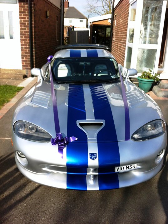 Got her out  - Page 1 - Vipers - PistonHeads - The image showcases a silver car with a distinctive blue stripe running down its center. The hood of the car is open and has a bow around it, suggesting it may be secured or perhaps part of a celebration or special event. The car is parked in a driveway with a brick wall in the background. The license plate of the vehicle is clearly visible, indicating it is a fully functional road vehicle. The overall scene gives a sense of readiness, as if the car is about to embark on a journey.