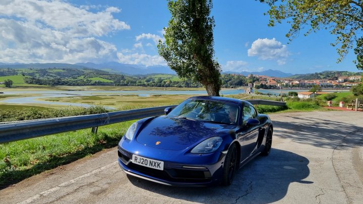 Pistonheads - The image features a sleek, blue sports car parked on the side of a road. The car is positioned facing towards the camera, allowing for a clear view of its design and features. It appears to be in good condition, with a glossy finish reflecting the sunlight.

In the background, there's a serene landscape with lush green trees and rolling hills under a blue sky dotted with clouds. The road on which the car is parked is lined with grass and trees, suggesting a rural or semi-rural setting.

There are also two individuals present in the scene, one standing close to the car while the other is slightly further away, both seemingly enjoying the outdoor environment. Their presence adds a touch of life to the otherwise inanimate scene.

The overall composition of the image suggests a peaceful, leisurely day out in nature, with the sports car serving as an impressive centerpiece amidst the tranquil surroundings.