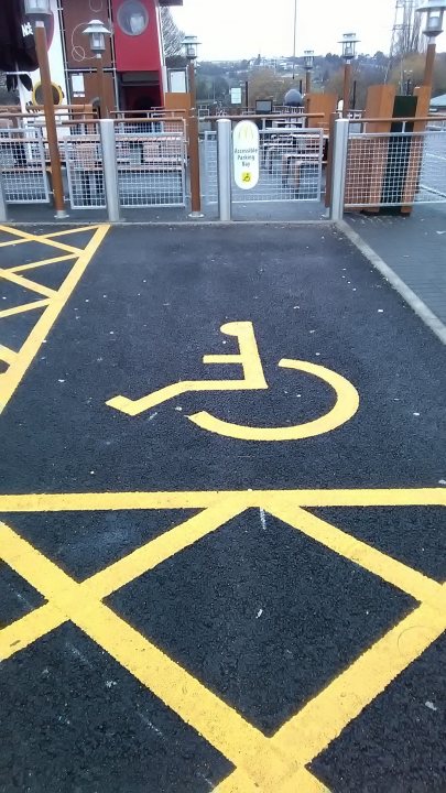 These pictures make my teeth itch - Page 288 - The Lounge - PistonHeads UK - The image shows a view of a parking lot with a marked disabled parking space. A yellow handicap symbol is prominently displayed on the pavement in front of the parking spot, indicating it's designated for people with disabilities. The background reveals a cloudy sky and what appears to be a body of water or a large puddle, reflecting the overcast conditions.