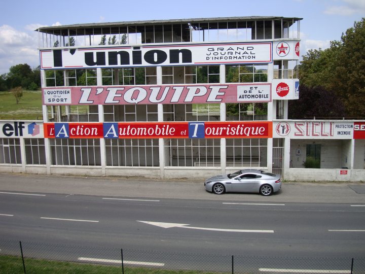 French trip - Page 1 - Aston Martin - PistonHeads - The image shows a silver car parked on the side of a street next to a building with large signage. The building has a prominent signboard at the top with the words "L'UNION TRANSPORT D'INFORMATION" and "GESTION AUTOMOBILE TRANSPORT". Below this, other signs include "L'ACTION AUTOMOBILE", "TOURISTIQUE", and other words that are partially obscured. The setting appears to be in an urban area during the daytime. The car has alloy wheels and is seated against some grass, adjacent to a paved area with a fence visible in the foreground.