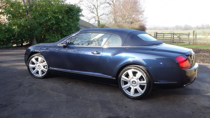 A classic car parked in a parking lot - Pistonheads - The image showcases a sleek, blue convertible car parked on a driveway. The vehicle has a hardtop that's currently retracted, revealing the soft-top interior. The car features large, silver-colored wheels that contrast with the dark exterior. In the background, you can see lush greenery, suggesting a suburban setting. A wooden fence is visible at the edge of the driveway, adding a rustic charm to the scene.