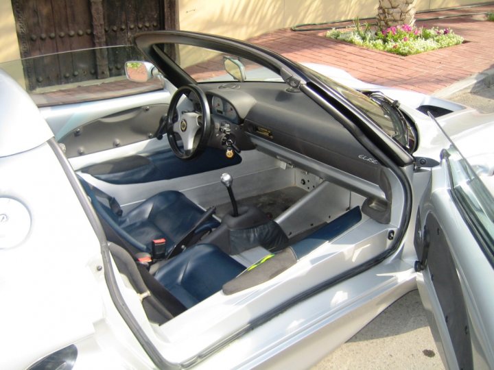 A car is parked on the side of the road - Pistonheads - The image presents an open, driver's side door of a convertible car. The interior, visible through the door frame, features a black steering wheel and blue leather seats. Two seats are noticeable with the driver's seat being closer and placed next to a U-shaped center console filled with controls and a candy-colored object. The car is parked in front of a building made of bricks, and there are various potted plants nearby. The sky appears clear, hinting at good weather conditions.