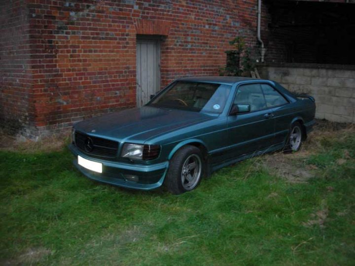 A long shot, but anyone know what happened to this SEC?  - Page 1 - Mercedes - PistonHeads - The image displays a vehicle parked in front of a brick wall with an open door, possibly indicating it has been abandoned or left incomplete. The car has a dark body and appears to be an older model, given its boxy shape and conspicuous proportions. The grasses surrounding the vehicle are tall, suggesting the car has been parked in this location for a considerable length of time, as the growth is quite substantial. The setting suggests a neglected or unused space, perhaps indicating the car's owner or age.