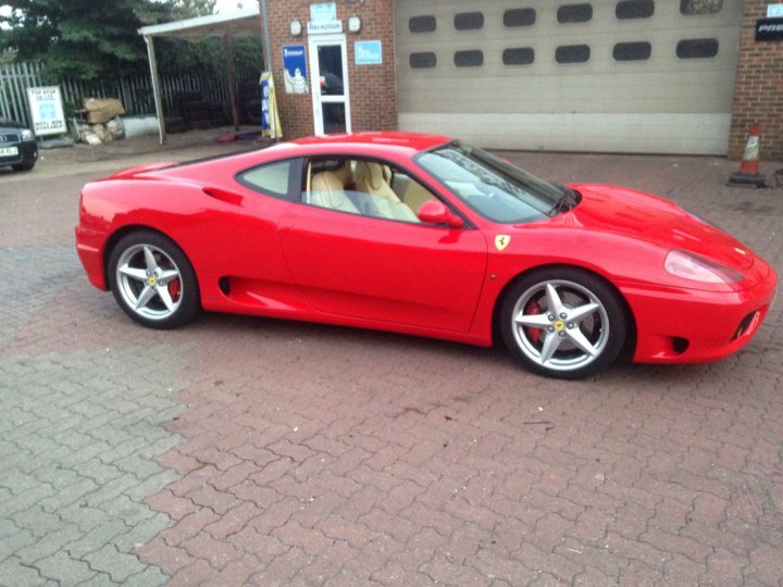 Hi... - Page 1 - Ferrari V8 - PistonHeads - The image shows a vibrant red Ferrari sports car parked on a brick pavement. The car is positioned at an angle, with its front facing towards the left side of the image. The Ferrari's sleek design and the iconic yellow Prancing Horse emblem are visible. In the background, there is a building covered in black metal shutters and a white door, which appears to be the entrance to a garage or workshop. There are also a few other vehicles visible in the top left corner of the image, suggesting this might be a commercial or parking area.