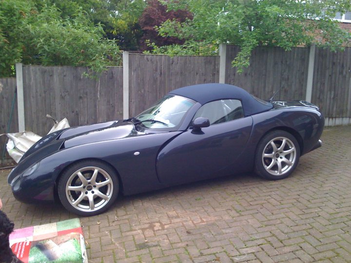 Photoshop wire wheels. - Page 1 - General Gassing - PistonHeads - The image presents a sleek black sports car parked on a brick driveway. The car is facing towards the left side of the image, allowing us to clearly see its aerodynamic design and alloy wheels. The setting appears to be a residential area, given the presence of a wooden fence to the left of the car and some shrubs in the vicinity. The car's position on the driveway suggests it's stationed at someone's residence, parked parallel to the curved fence line. The overall scene indicates a tranquil, suburban atmosphere.