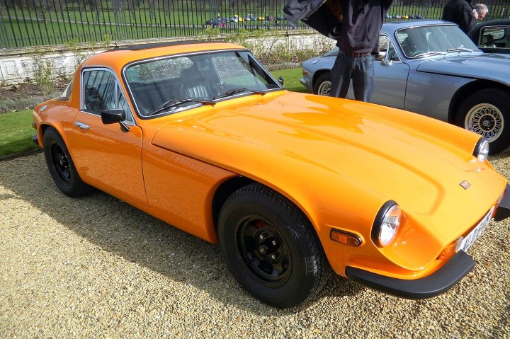 Early TVR Pictures - Page 90 - Classics - PistonHeads - The image captures a tranquil scene, dominated by a group of vintage cars parked on a gravel driveway, bathed in the soft glow of early morning light. The most striking of these vehicles is a vibrant orange sports car, its sleek, iconic shape standing out against the more subdued offerings behind it. The sports car is further enhanced by its contrasting black roof and matching black wheels, a testament to classic motoring aesthetics. A man, partially visible on the side, appears to be appreciating the vintage cars, adding a sense of human interest to the scene. The backdrop of the image reveals a lush, verdant garden, dotted with trees under a clear sky, providing a natural, serene setting for this tableau of old-world elegance.