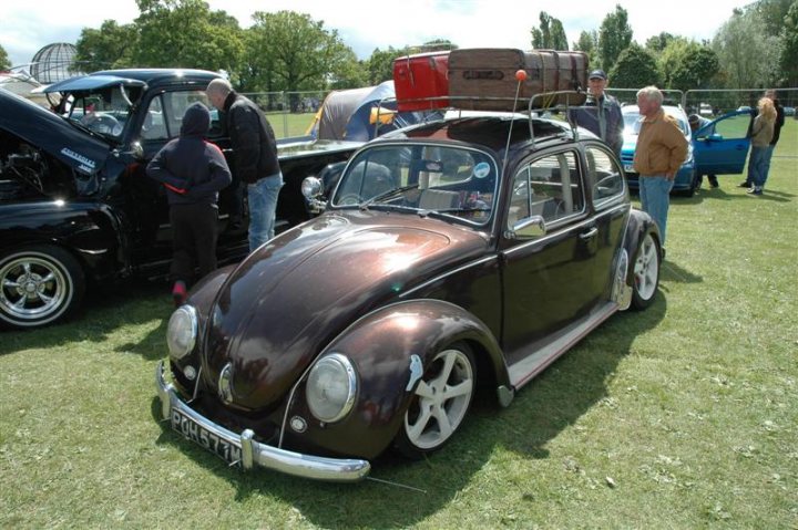 Fins and Chrome - Page 1 - North East - PistonHeads - The image showcases a classic Beetle car parked on grass. The car is black and well-maintained, with a vintage design that adds a touch of nostalgia. A distinctive feature of the car is a surfboard tied to its roof, suggesting it might be used for beach trips. 

In the background, there are several other vintage cars parked in a row, creating a space that seems to be a gathering spot for classic vehicle enthusiasts. The grassy setting and the casual arrangement of the cars suggest a relaxed and leisurely atmosphere, likely a car show or a meetup for vintage car owners.