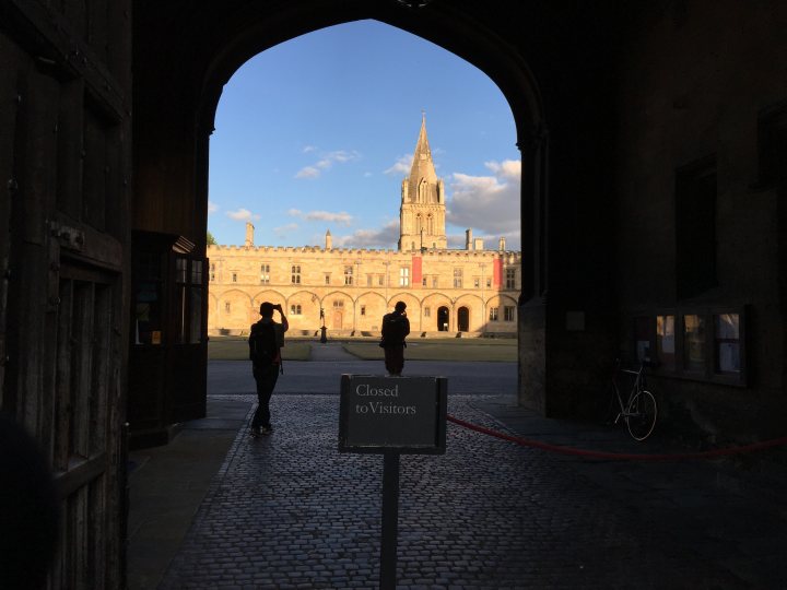 The "Photos From Today's Ride" thread... - Page 171 - Pedal Powered - PistonHeads - The image showcases a medieval-style stone building with a white-domed tower that pierces the sky in the background. A wooden gate with reinforced metal details is slightly ajar, giving a glimpse into the building. Behind the gate, two individuals can be seen walking away on a cobblestone pathway, adding a sense of life and movement to the scene. The setting appears to be a historic or university campus, as evidenced by the sign to the left in the foreground. The overall atmosphere is one of quiet serenity, highlighting the architectural beauty and historical significance of the building.