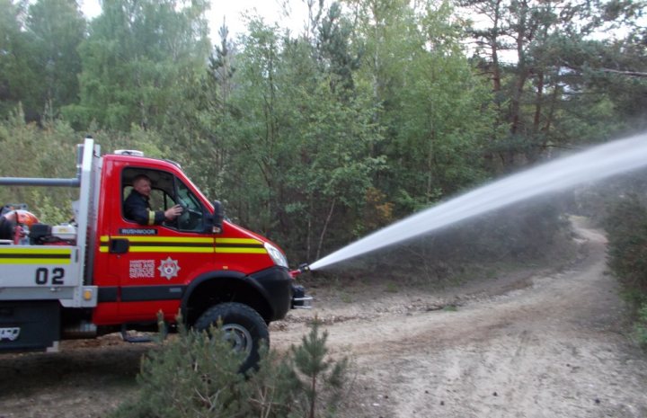 The "Photos From Today's Ride" thread... - Page 250 - Pedal Powered - PistonHeads - The image depicts a wilderness scene where a firefighting truck is actively engaged in controlling a river of water. The truck, parked on a dirt road, has its door open with a person inside. A hose attached to the side of the truck extends over the vehicle, directed at a river of water running beside the road. The person inside the truck seems to be involved in managing the control of water. The surrounding area is rich in trees, showing a dense forest environment. The scene suggests an emergency situation or a water pumping operation taking place in a remote, natural setting.