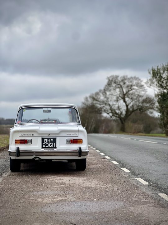 Pistonheads - The image presents a serene rural scene. Dominating the center of the frame is an old, light-colored car parked on the side of a road, which cuts across the image from left to right. The car's vintage design and the absence of modern vehicles suggest a timeless quality to the photo.

The sky overhead is filled with clouds, casting a soft, diffused light over the scene. The road itself appears well-maintained, hinting at a well-traveled route. 

On the right side of the image, there's a lone tree standing tall against the backdrop of the sky. Its presence adds a touch of nature to the otherwise man-made elements in the scene. The overall composition of the image creates a sense of tranquility and solitude.