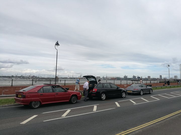 My bodged Citroen BX 16v - Page 12 - Readers' Cars - PistonHeads - The image depicts a scene on a grey and cloudy day by the sea. Several cars are parked along the road, which is separated from a sandy beach with railing. The cars range from a red hatchback, a black sedan, and a silver coupe. Some people are standing near the open trunks of the cars, possibly loading or unloading items. The distant view includes a body of water, a bridge, and a city skyline, suggesting that they are near a coastal urban area.