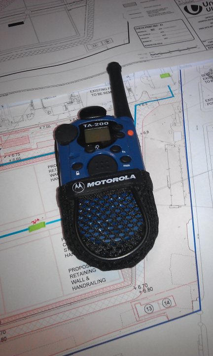 Two way radio? - Page 1 - Aston Martin - PistonHeads - The image features a blue Motorola walkie-talkie prominently placed on top of a blueprint. The walkie-talkie, characterized by its antenna and a directional microphone, is the main object in the image. The blue print beneath it contains lines and text, suggesting it could be a technical or architectural drawing. The document has a ruler drawn on it, indicating measurements are being taken or recorded. The contrast between the man-made object and the technical drawing hints at a construction or engineering context.