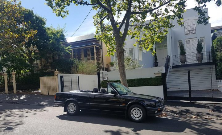 Lovely Cars: Interesting, Classic, Retro, Barge 5-10k - Page 60 - General Gassing - PistonHeads - The image depicts a quiet residential street on a sunny day. At the center of the scene is a black convertible car parked on the right side of the street. The car is sleek and shiny, suggesting it is somewhat new. Behind the car, a white privacy fence can be seen along with white walls of a house, indicating a well-maintained property. The house is framed by lush green trees that line the street, adding to the serene and peaceful ambiance.