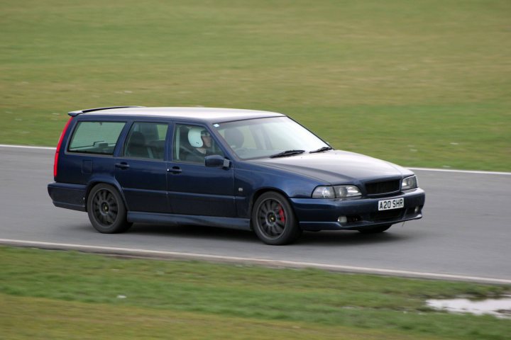 Track day at Snetterton Friday 15th February - Page 1 - East Anglia - PistonHeads - The image captures a scene on a curvy road. A predominantly blue A40 Quattro Audi, sleek and well-maintained, is the primary subject. It's in motion, its nose pointed towards the right side of the image, suggesting it's turning or navigating a curve. A number of smaller, indistinct objects, possibly cars or signs, can be seen in the background. The grassy embankment adjacent to the road leads to a small body of water, adding a natural element to the scene.