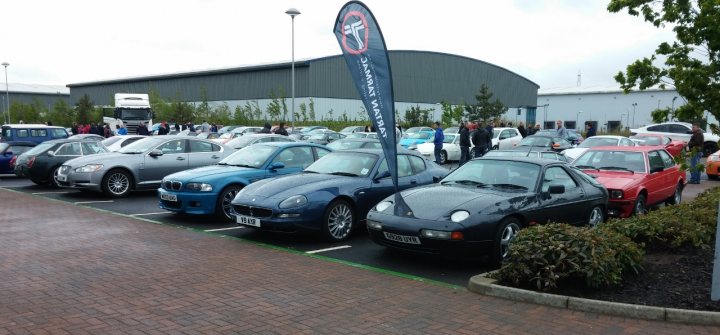 A bunch of cars that are parked in a lot - Pistonheads - The image captures a lively scene in a parking lot, where a multitude of cars are neatly parked in a row. The cars vary in color and model, adding a vibrant mosaic to the setting. A group of people, presumably the owners or spectators, are scattered around the area, some standing near their cars, engaged in conversation, while others are merely passing by. In the background, a large building extends into the horizon, perhaps indicating the proximity of a convention or auto show event. Four banner stands rise amidst the parked cars, each sign displaying a prominent logo, suggesting that they are likely sponsors or exhibitors at the event. Planted along the border of the lot are bushes, adding a touch of greenery to the surroundings.