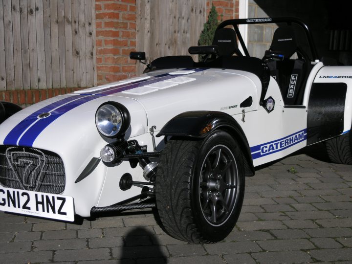 What colour would you go for ? - Page 2 - Caterham - PistonHeads - The image features a sporty, one-seat motorsport car parked on a brick surface. It's painted white with accents of blue and black on the front and sides, and it has numbered tags. The car is characterized by large frontal air vents, enhanced fenders, and a sleek design that looks futuristic. Behind the car, there's a brick wall with a wire mesh. The vehicle is positioned at a slight angle to the camera, highlighting its profile.