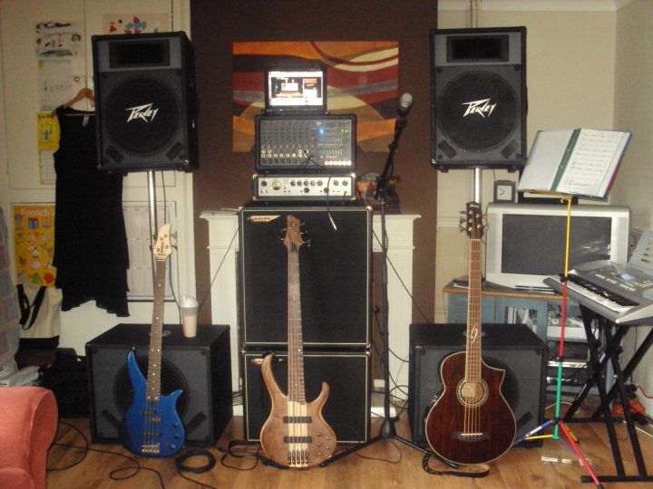 Pistonheads Musicians - The image is a bustling scene filled with an array of electronic music equipment and guitars. A black bass guitar stands prominently in the foreground, alongside a purple electric guitar. An acoustic guitar leans comfortably to the side, its pick guard visible. Two large speakers tower on either side of the room, their logos spelling "TASY". Atop these speakers is a television set, turned off and awaiting its next performance. On the right, a keyboard setup is visible, hinting at the possibilities of this music studio. The floor is carpeted in a dark hue, contrasting with the white walls and clean lines of the room.