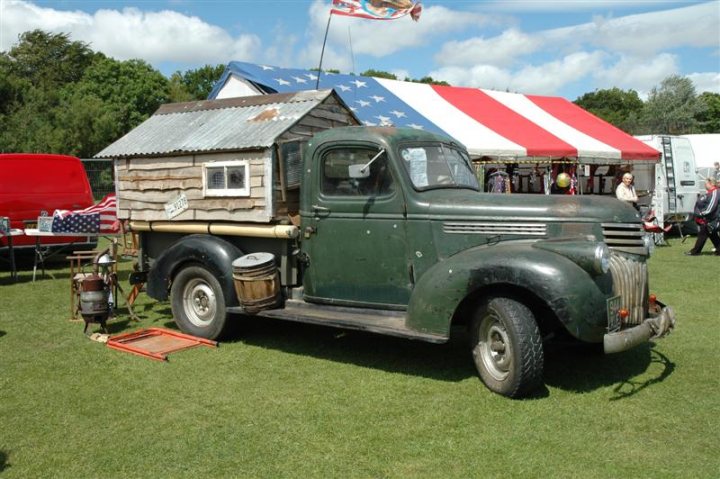 Fins and Chrome - Page 1 - North East - PistonHeads - The image shows an older green truck with a rusted, wooden roof that resembles a wagon train cabin. The truck is parked on grass and it prominently displays the American flag pattern on its roof and an American flag blowing in the breeze. The tent in the background has a similar flag pattern, but here it is the national flag, which could indicate the location or context of the gathering, possibly a patriotic celebration or a promotional event for an American product or company. There are some people visible in the distant background, possibly attendees or staff, but the exact nature of the event isn't clear from the image.