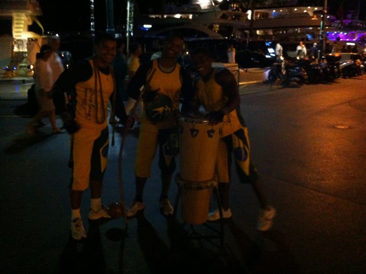 A group of people walking down a street - Pistonheads - The image depicts a group of four men standing together on a street at night, smiling at the camera. They are wearing matching yellow and green tracksuits, holding a large yellow percussion instrument between them. The men's position and the instrument they are holding together suggest they may be musicians performing. The setting is outdoors with other people visible in the background, near water, indicating a lively social event or gathering.