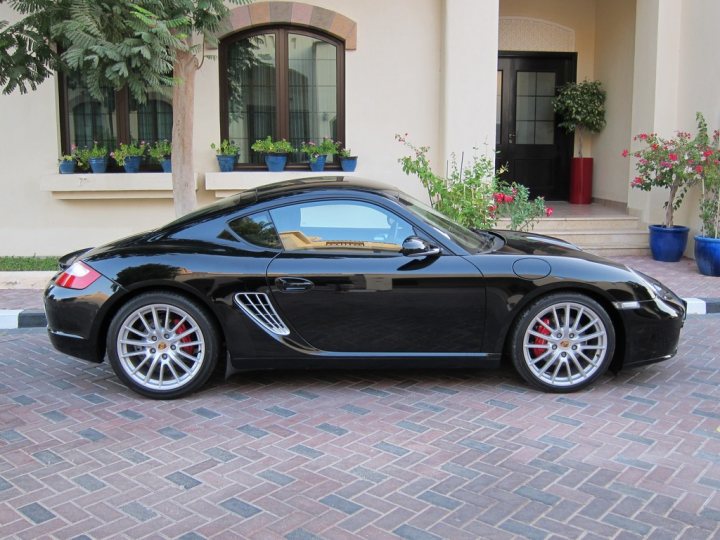 Would a Manual Boxster/Black Leather be Financial Suicide!? - Page 1 - Middle East - PistonHeads - This image features a classic black car parked on a brick driveway. The car has distinctive red trim and silver accents, suggesting it is a luxury or sports vehicle. The brick paving has a clean, well-maintained appearance, and there are planters with various plants and flowers on either side of the entrance, adding a touch of greenery to the scene. The building in the background has columns and a window box, suggesting a traditional architectural style.