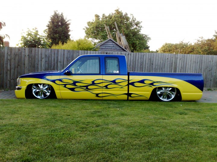 Pistonheads Truck Chevy - The image features a pickup truck with a vibrant flame paint job on its sides, that appears to be stationary on grass. The vehicle exhibits a predominantly blue and yellow color scheme, with the flames painted in yellow against a blue background. It possesses large, chrome wheels and a sleek body style. In the background, there is a wooded area and a fence with privacy panels, suggesting a residential setting. The sky is overcast, casting muted light on the scene.