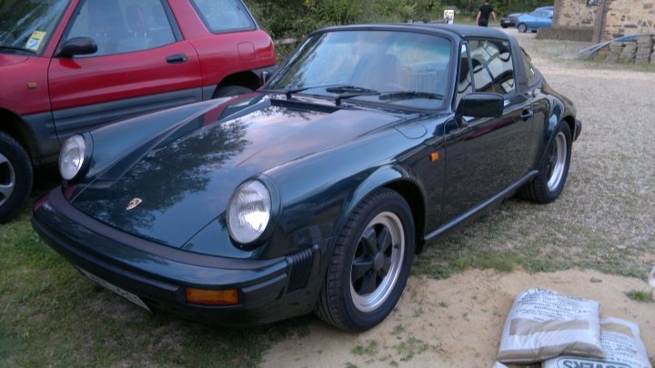 A car is parked in a parking lot - Pistonheads - The image shows a black classic car parked on a gravel driveway. The car has a glossy finish and features a visible, round silver emblem on the center of the hood. It has a two-door design, with a convertible top that appears to be closed. The car is located in front of another vehicle, which is a red SUV, suggesting the scene is taken in a residential area with parked cars. The lighting in the photo indicates it might be either early morning or late afternoon, as there are shadows visible on the ground. There are no texts visible in the image.