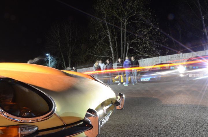 Essex Pistonheads March - The image shows a group of people gathered around a vintage car at night. The car appears to be an older-model sports car, possibly a Porsche based on the distinctive front design. The car is beige or light gold in color, and it is parked on a street or parking lot. The lighting in the photograph suggests that the camera flash captured the moment, creating a stark contrast between the illuminated car and the surrounding darkness. The people, likely friends or enthusiasts, are standing and appear to be speaking with each other, their bodies visible as silhouettes against the light from the car. The overall mood of the image is casual and relaxed, with a clear focus on the car and the gathering of people around it.