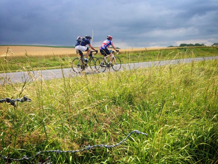 The "Photos From Today's Ride" thread... - Page 150 - Pedal Powered - PistonHeads - This image captures a dynamic scene of two cyclists energetically pedaling down a country road. They are both wearing helmets for safety, while the rider on the left sports a blue jersey paired with a white shorts and their bike bears both British and American flags. 

The setting is a picturesque rural landscape with tall, unkempt grasses bordering the road on both sides, and a wire fence can be spotted in the foreground, suggesting a boundary enclosing the area. The sky in the background is filled with clouds, adding a sense of depth and distance to the image. 

The focus of the image is the cyclists and their movement, conveying a sense of speed and adventure as they traverse the green country road. The colors and elements in the image work together to create an engaging and lively scene.