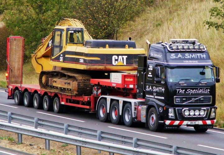 RE: Mr Big Stuff News: Go Green With Big Trucks - Page 4 - General Gassing - PistonHeads - The image captures a dynamic scene on a highway where a large hauling truck is towing an even larger piece of cat equipment. The truck is black, while the backhoe on its bed exhibits yellow and orange hues. The vehicles are in motion, and the road ahead is flanked by metal barriers and lush green grass. The inscription "S.A.Smith The Specialist" is visible, indicating the truck company that specializes in equipment transportation.