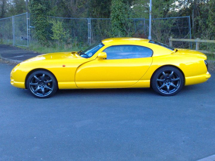 Considering returning to the TVR fold.. - Page 3 - Tamora, T350 & Sagaris - PistonHeads - The image captures a bright yellow sports car gracefully parked on a driveway. The car's sleek design and black tire rims contrast beautifully with the yellow body. It's positioned in front of a chain-link fence, which, along with the vegetation in the background, adds a sense of depth to the scene. The driveway itself is paved, providing a reflective surface that mirrors the car's vibrant color and enhances the striking visual effect. The overall composition suggests a peaceful, suburban setting, where the car is likely waiting for its proud owner to return.