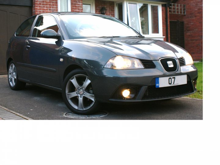 Ibiza Whip Sport Pistonheads Seat - The image features a modern, dark-colored hatchback parked on a gray paved driveway. The vehicle's design is sleek and contemporary, with visible creases in the bodywork, and it is equipped with a spare tire mounted on the trunk hatch. The car has a distinctive front grille, featuring the emblem of the manufacturer, a European license plate displaying the number "07," and headlights that are currently illuminated, suggesting that the photo was taken in the evening or at night. The house in the background appears to be of brick construction, and there are clear skies above, indicating good weather.