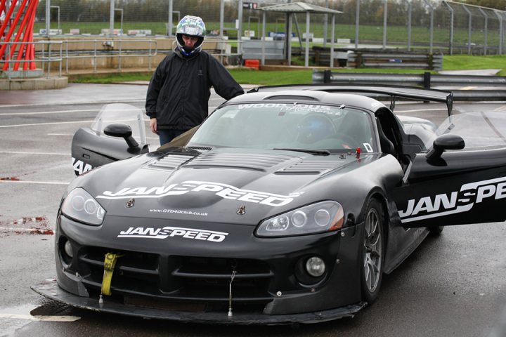 Pistonheads - This image shows a person standing next to a parked black racing car. The car is impressive, with a large spoiler and detailed bodywork. The person is dressed in black and is also wearing a helmet, suggesting they might be a driver or a driver's assistant. The scene appears to be set in a garage or assembly area. The focus of the image is the car, with the person providing a sense of scale to emphasize the size and design of the vehicle.