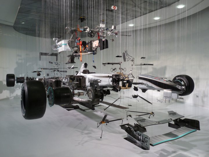 A bunch of luggage that are in a museum - Pistonheads - This image displays an elaborate and complex installation of apparent mechanical components, related to a car, suspended from a curved ceiling. The arrangement of these components creates an overhead view that resembles an engine block or a series of interconnected parts against a backdrop that could signal an evolving or fractal-like design. The setting suggests a modern, possibly architectural art space, with its minimalistic background and the symmetry of the components. The composition is dense and yet detailed, where each piece of metal has its own distinct shape and form.