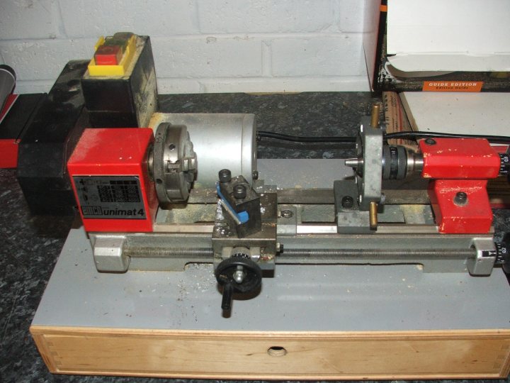 Change Speed Pistonheads Lathe - The image shows a well-used industrial machine with various parts and components. It appears to be a drill press, as evidenced by the circular drill and the adjacent frame. The machine is situated on a concrete floor. There's no person in the frame; the focus is entirely on the machine.