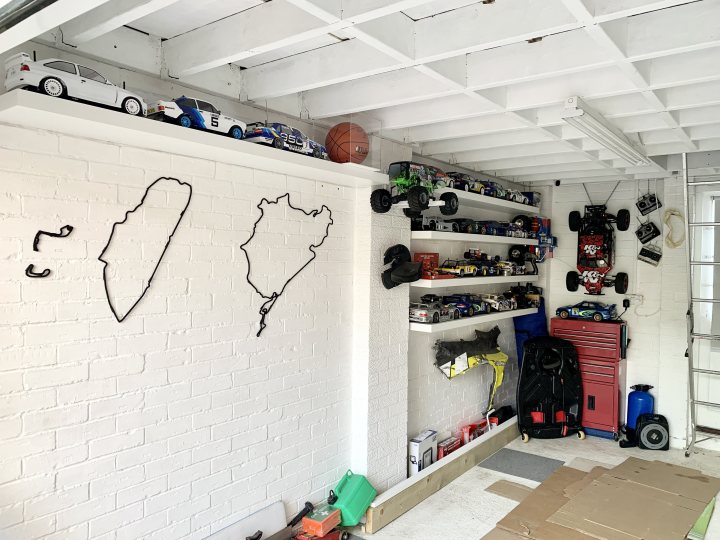 Show us your RC - Page 25 - Scale Models - PistonHeads UK - The image shows a room that appears to be a garage or storage area. On one side, there's a whiteboard with two hand-drawn maps on it; one of Israel and the other of Europe. Above this whiteboard, a row of shelves holds various objects like cars, trucks, and toy racecars. There is also a small sports ball placed among these items. The floor is covered by a black and white checkered pattern. On the opposite side, there are additional shelves holding more toys, including more cars and trucks, as well as other miscellaneous items. A cardboard box and a couple of red and blue suitcases can be seen in the middle of the room. The walls of the room are partially covered with white bricks and wooden planks, giving it an industrial look. There is no visible text in the image that provides additional context or information.