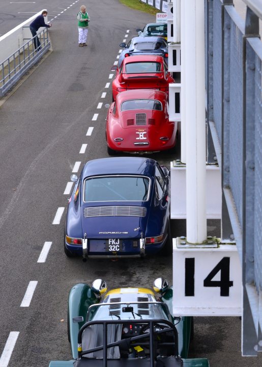 Robert Barrie Track day 24th Feb - photos - Page 1 - Goodwood Events - PistonHeads - The image showcases a bustling car show, with a line of vintage and sports cars neatly parked on either side of a white road with white markings. A variety of classic car models, from convertibles to racers, can be seen. Their vivid colors range from red and blue to green and yellow, adding a vibrant touch to the scene. The cars are facing the same direction, possibly hinting at a start line or a track-side setting for an ongoing car show or race.